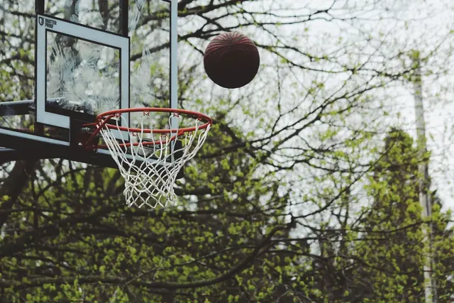 A basketball in the air above an outdoor hoop among trees