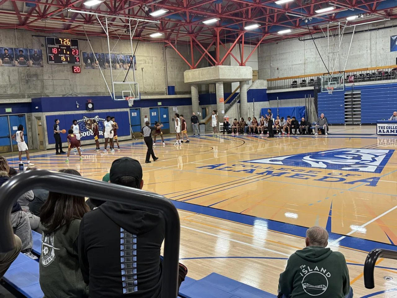 A College of Alameda men’s basketball game in the Cougars’ gym, with players at the free-throw line, both benches and a scoreboard