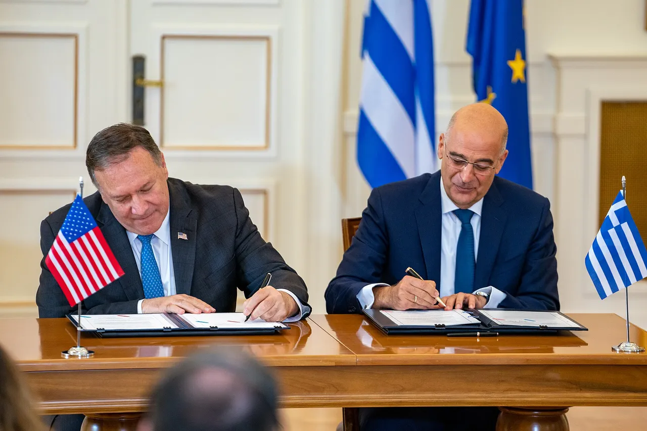 Two officials sit at a wooden table and sign documents with pens during a formal agreement signing ceremony.