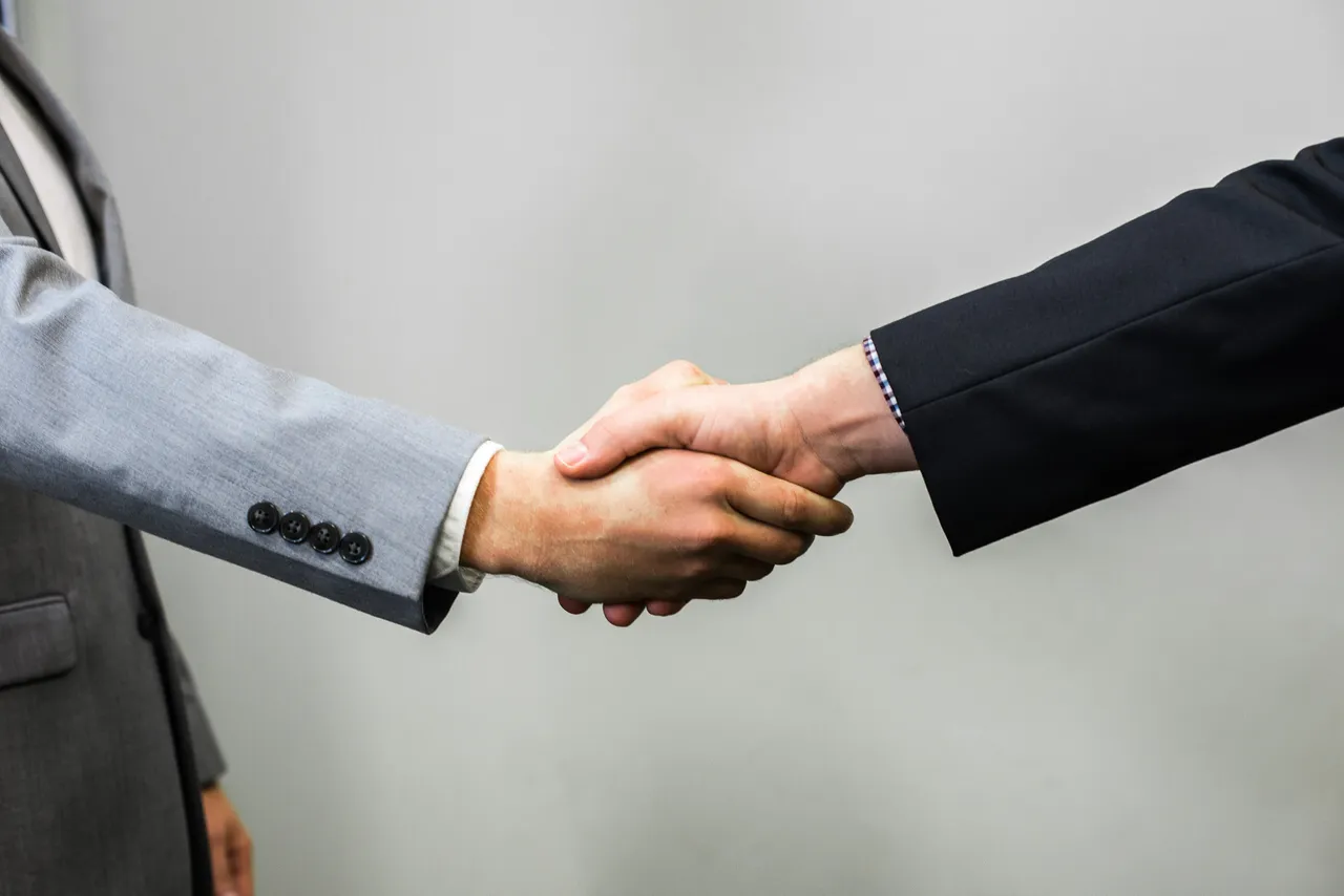 Two men in business suits shake hands against a plain gray wall.
