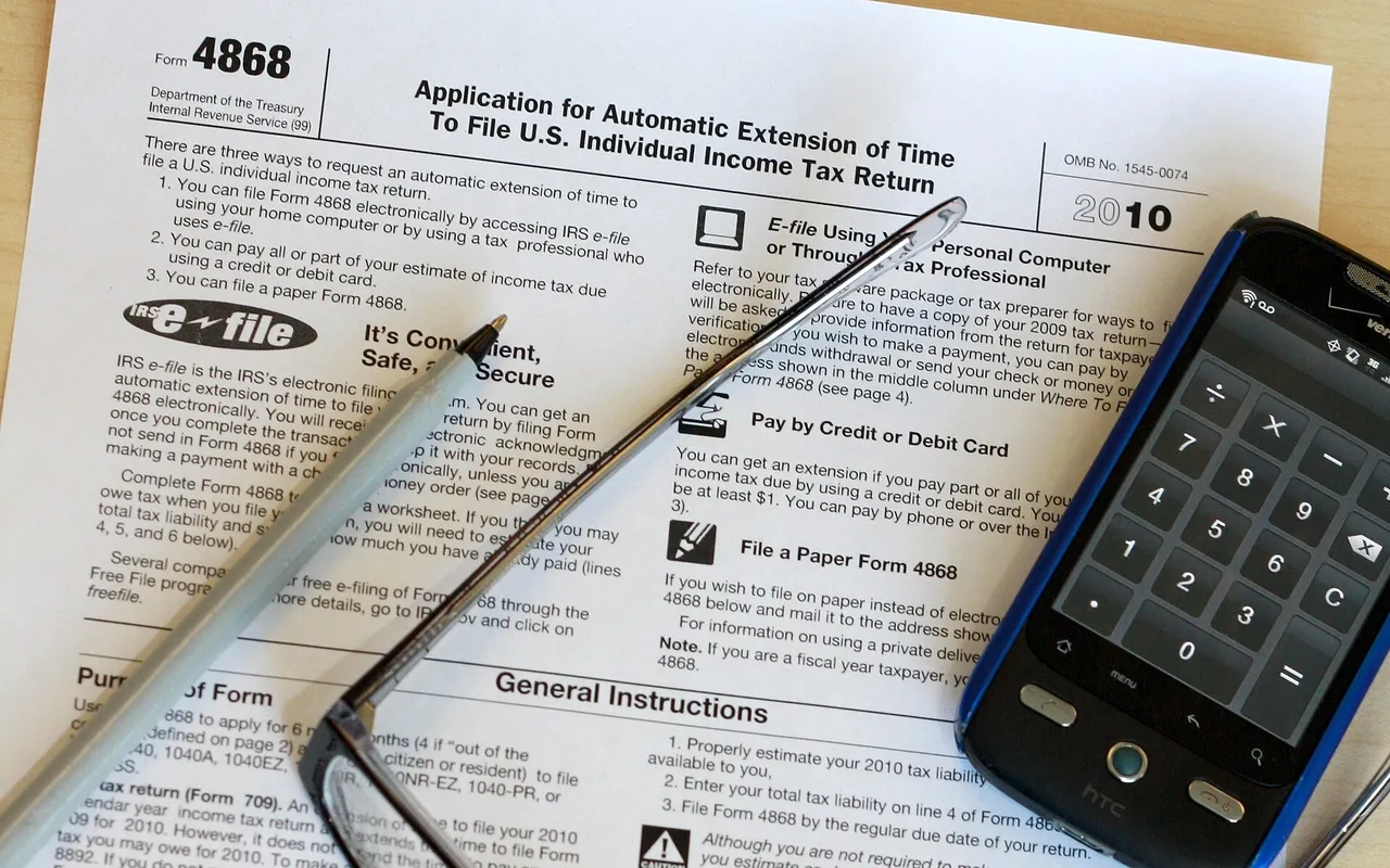 An IRS Form 4868 tax form on a desk with a pen, eyeglasses and a smartphone calculator on top.