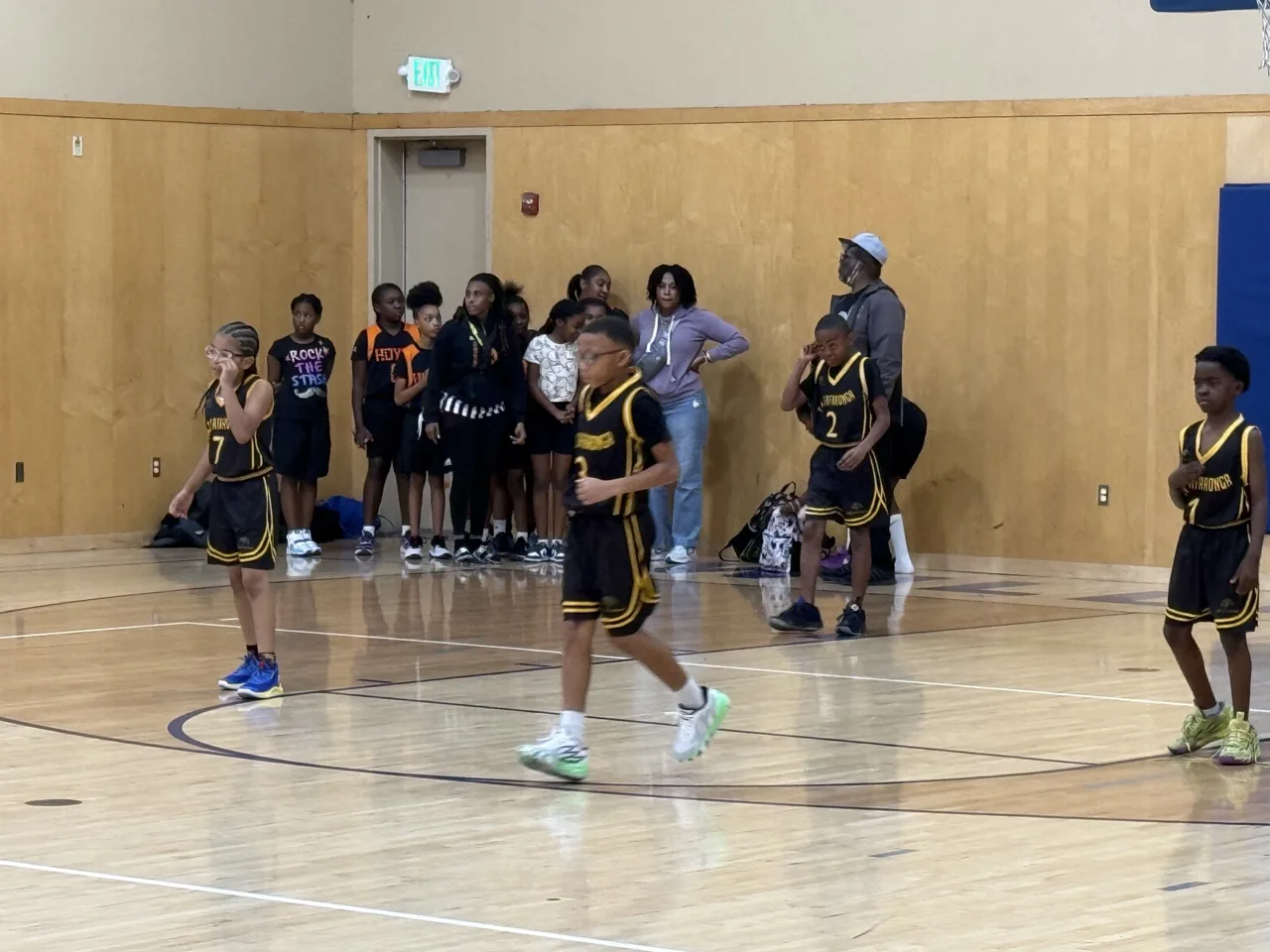 Youth players in black and gold uniforms spread out during a game in a recreation gym while the opposing team and a parent gather behind the baseline