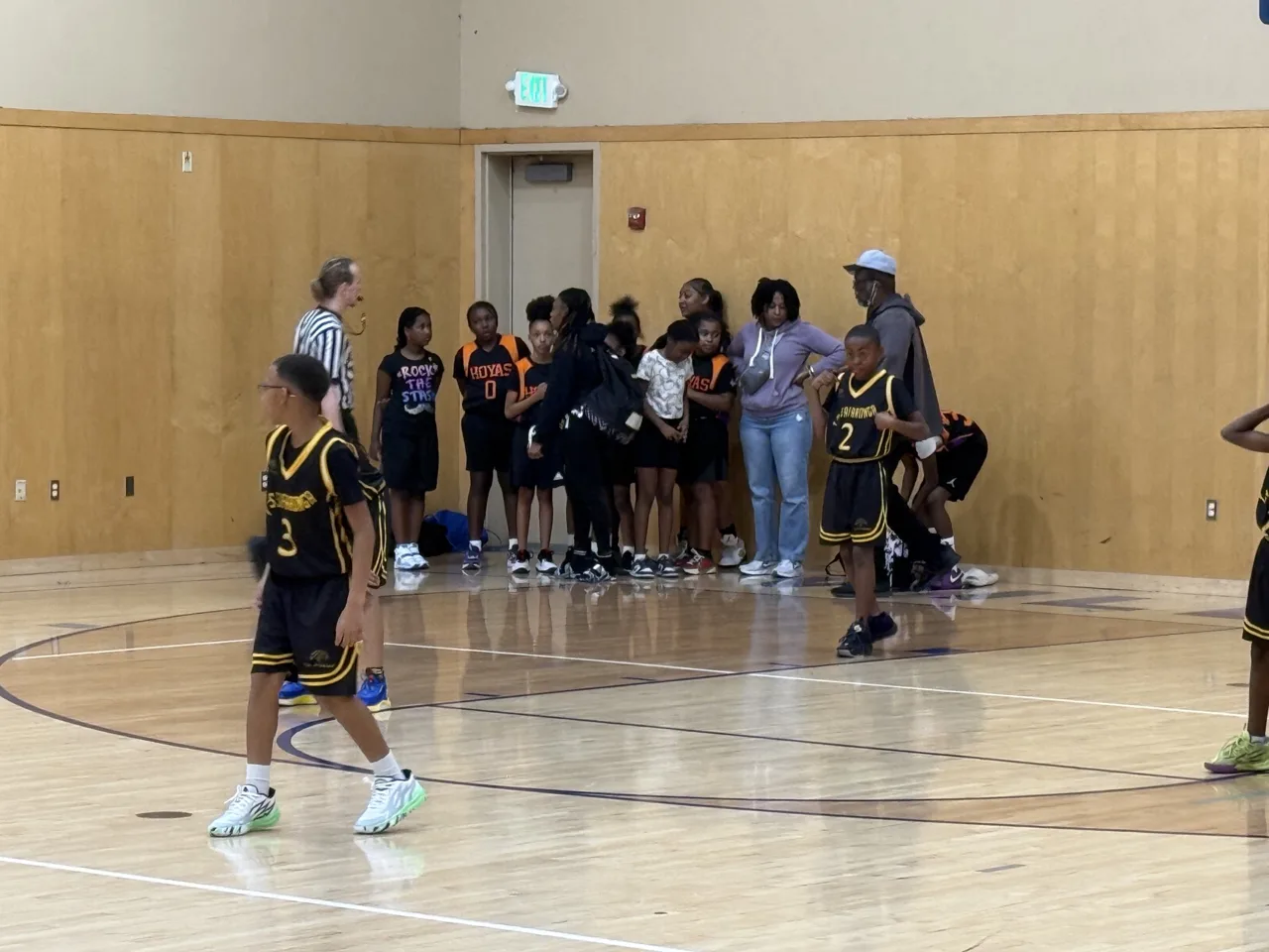 A referee and youth players in black and gold uniforms pause during a break in play in a recreation gym while the opposing team huddles by the wall