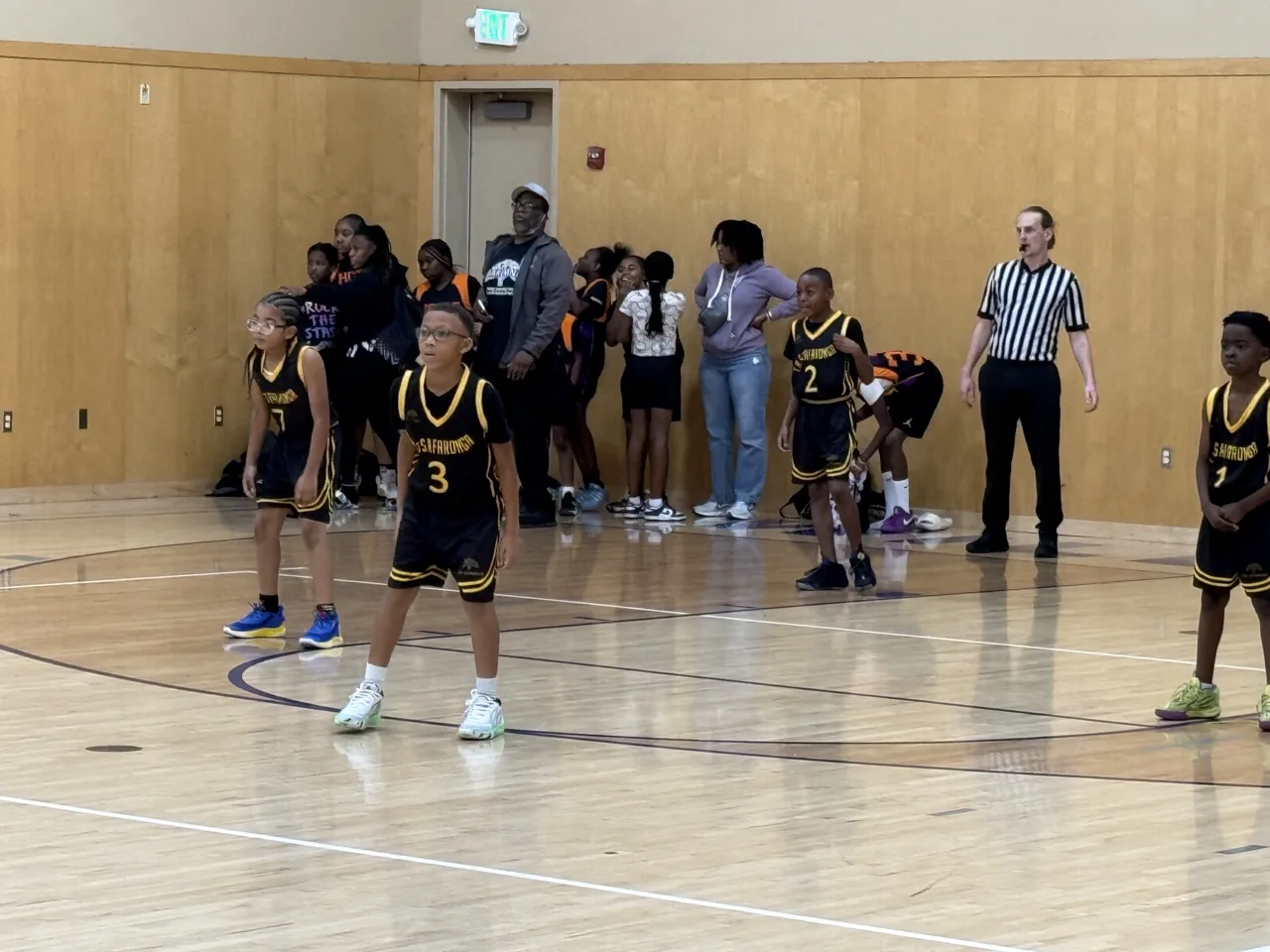 Youth players in black and gold uniforms wait for play to resume in a recreation gym, with a referee and the opposing bench behind them